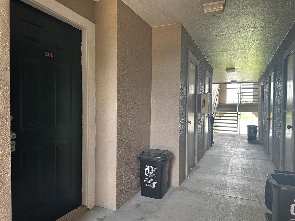 An outdoor apartment building hallway with a dark door labeled 205 and trash bins — 4209 WOODSTORKS WALK WAY UNIT 1205, LUTZ, FL