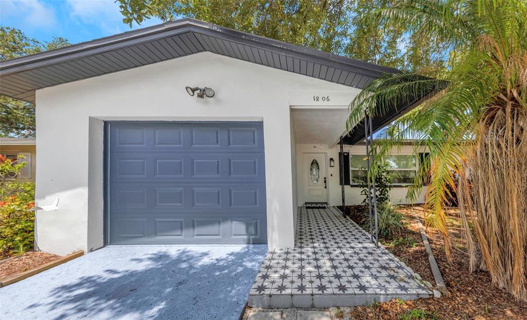 A white house exterior features a gray garage door and a tiled walkway to the entrance — 1806 VANCOUVER DRIVE, CLEARWATER, FL