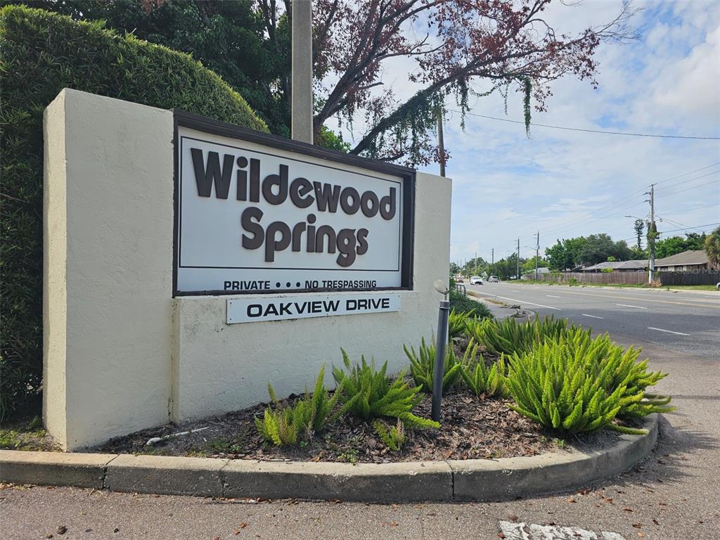 A sign for Wildewood Springs stands beside a road with landscaping in the foreground — 153 WILD PALM DRIVE UNIT 1610, BRADENTON, FL