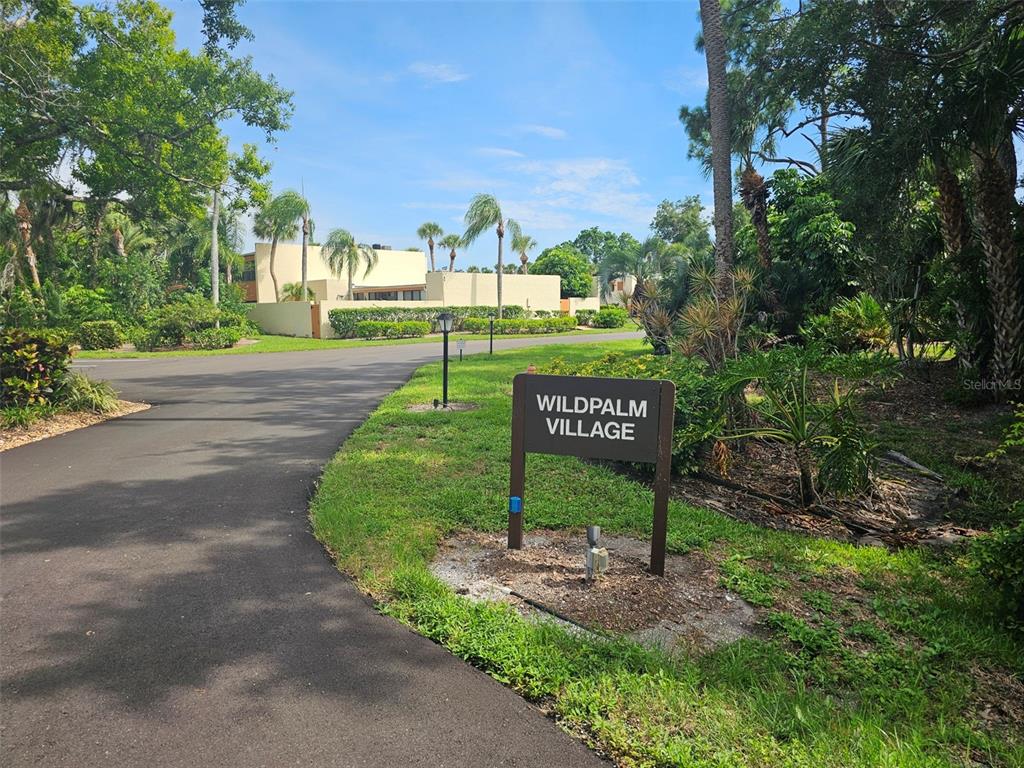 A paved road leads to a sign for Wildpalm Village surrounded by trees and landscaping — 153 WILD PALM DRIVE UNIT 1610, BRADENTON, FL