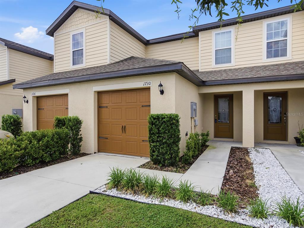 A two-story townhouse exterior with tan siding, a brown garage door, and a concrete walkway — 1730 WYNFORD CIRCLE, TAVARES, FL