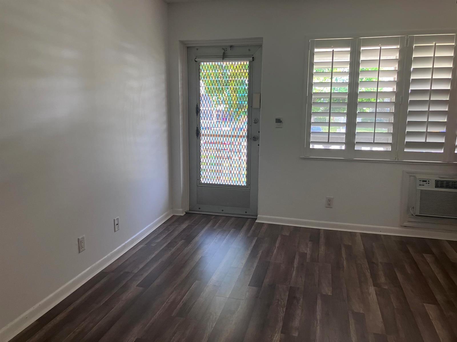 An empty room features wood-look flooring, white walls, a security screen door, and window shutters — 259 Sheffield K # 259, West Palm Beach FL 33417, WEST PALM BEACH, FL