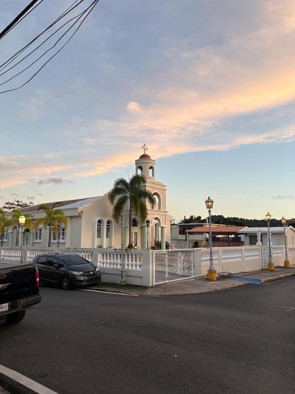 A light-colored church with a bell tower stands behind a white fence at sunset — 348 KM 9.1 C. ENRIQUE PAGAN- POBLADO ROSARIO, SAN GERMAN, PR