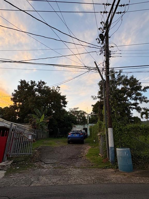 A blue car is parked on a gravel driveway leading to a house at sunset — 348 KM 9.1 C. ENRIQUE PAGAN- POBLADO ROSARIO, SAN GERMAN, PR