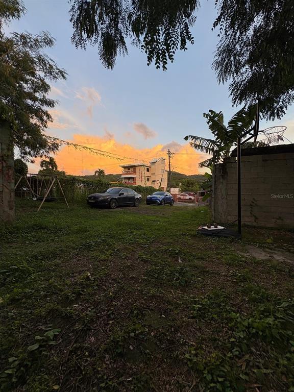 A grassy backyard features a basketball hoop, a swing set, and two parked cars at sunset — 348 KM 9.1 C. ENRIQUE PAGAN- POBLADO ROSARIO, SAN GERMAN, PR