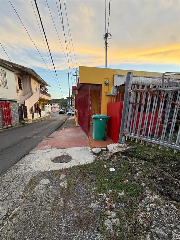 A yellow and red building sits along a street with a green trash bin outside — 348 KM 9.1 C. ENRIQUE PAGAN- POBLADO ROSARIO, SAN GERMAN, PR