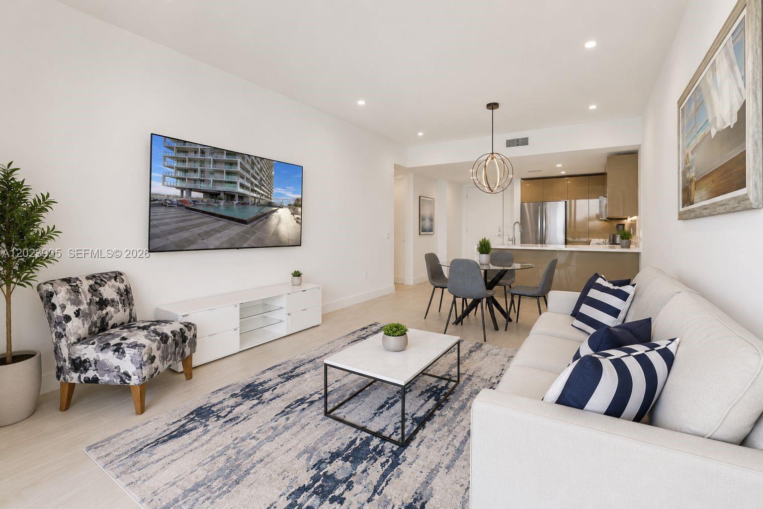 A living room features a white sofa, patterned armchair, television, and an adjacent dining area — 2000 Metropica Way # 1408, Sunrise FL 33323, SUNRISE, FL