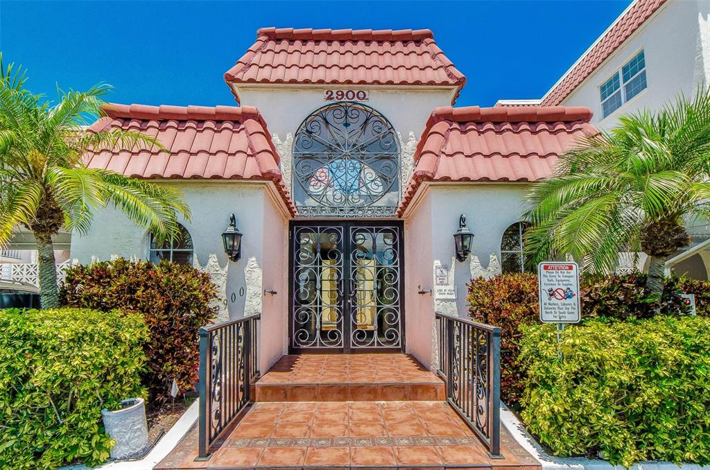 The exterior of a building entrance features a tiled staircase, decorative metal doors, and palm trees — 2900 GULF BOULEVARD UNIT 204, BELLEAIR BEACH, FL
