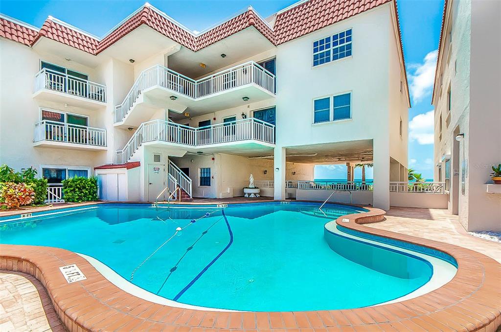 A multi-story apartment building with white exterior walls and balconies overlooks a curved swimming pool — 2900 GULF BOULEVARD UNIT 204, BELLEAIR BEACH, FL