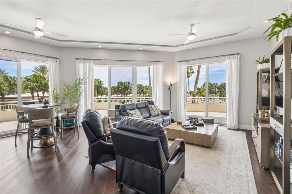 A living room with dark leather furniture, hardwood floors, and large windows overlooking palm trees — 1180 GULF BOULEVARD UNIT 101, CLEARWATER BEACH, FL