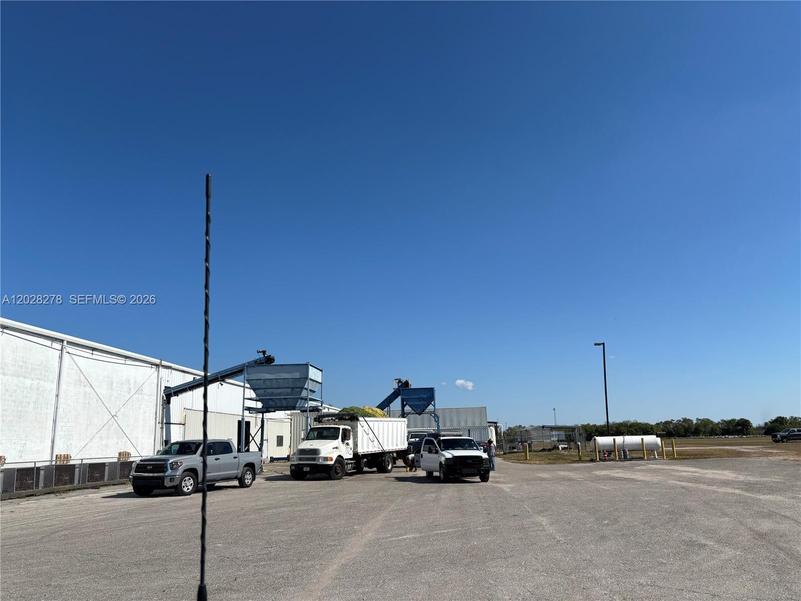 A paved industrial lot features a large white warehouse, trucks, and loading equipment under a clear sky — 15877 E SR 80, Clewiston FL 33440, CLEWISTON, FL