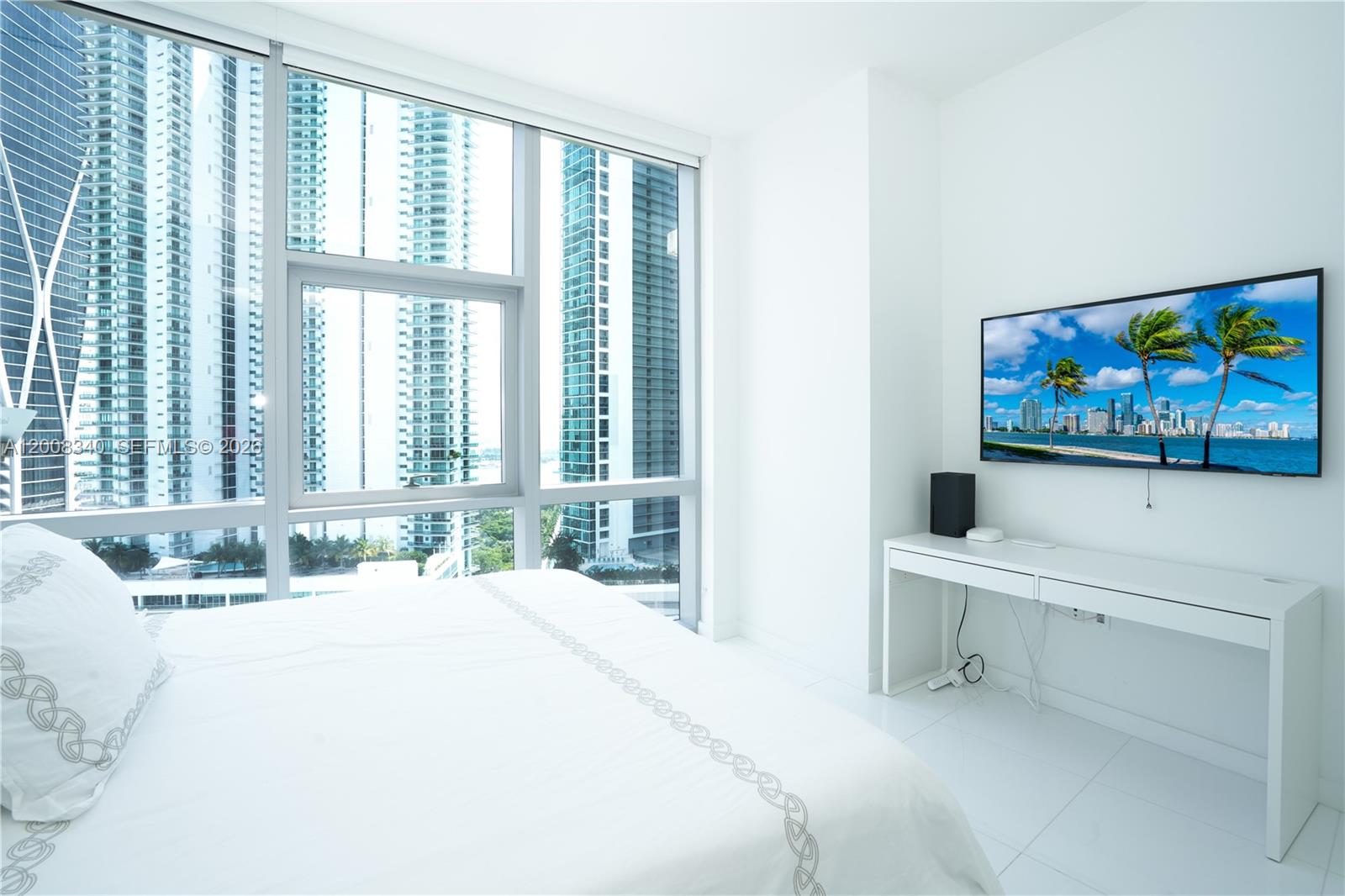 A bedroom with white furniture, a wall-mounted television, and floor-to-ceiling windows overlooking city skyscrapers — 851 NE 1st Ave # 2101, Miami FL 33132, MIAMI, FL