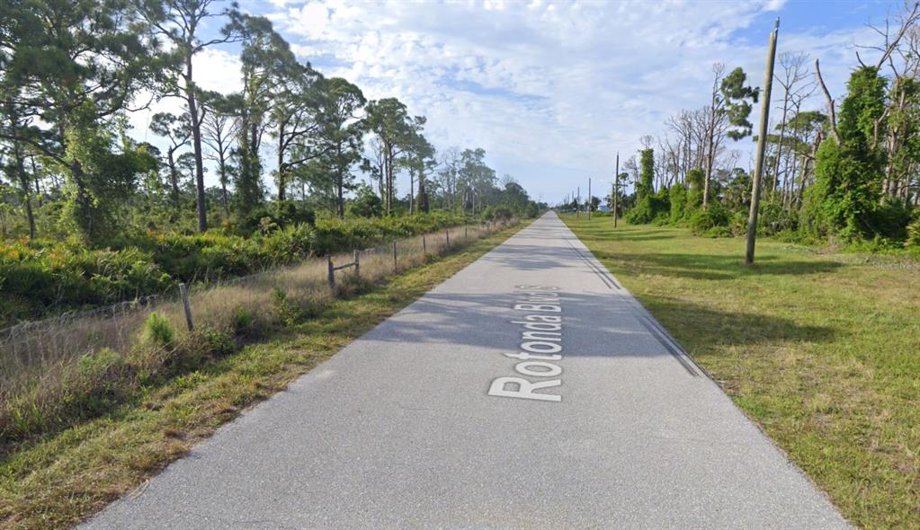 A paved road stretches into the distance, flanked by grassy shoulders and dense pine trees — 285 ROTONDA BOULEVARD S, ROTONDA WEST, FL
