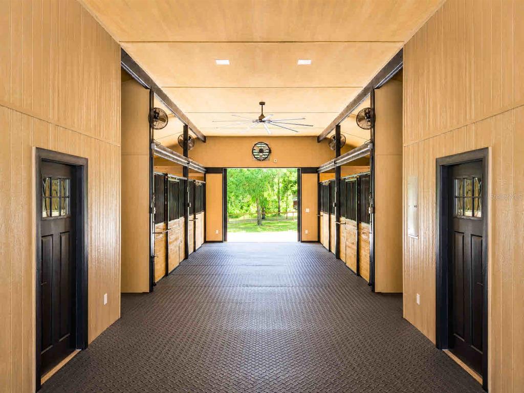 A horse barn aisle features rubber flooring, wood-paneled walls, black stall doors, and a ceiling fan — 196 NW 100TH AVENUE, OCALA, FL