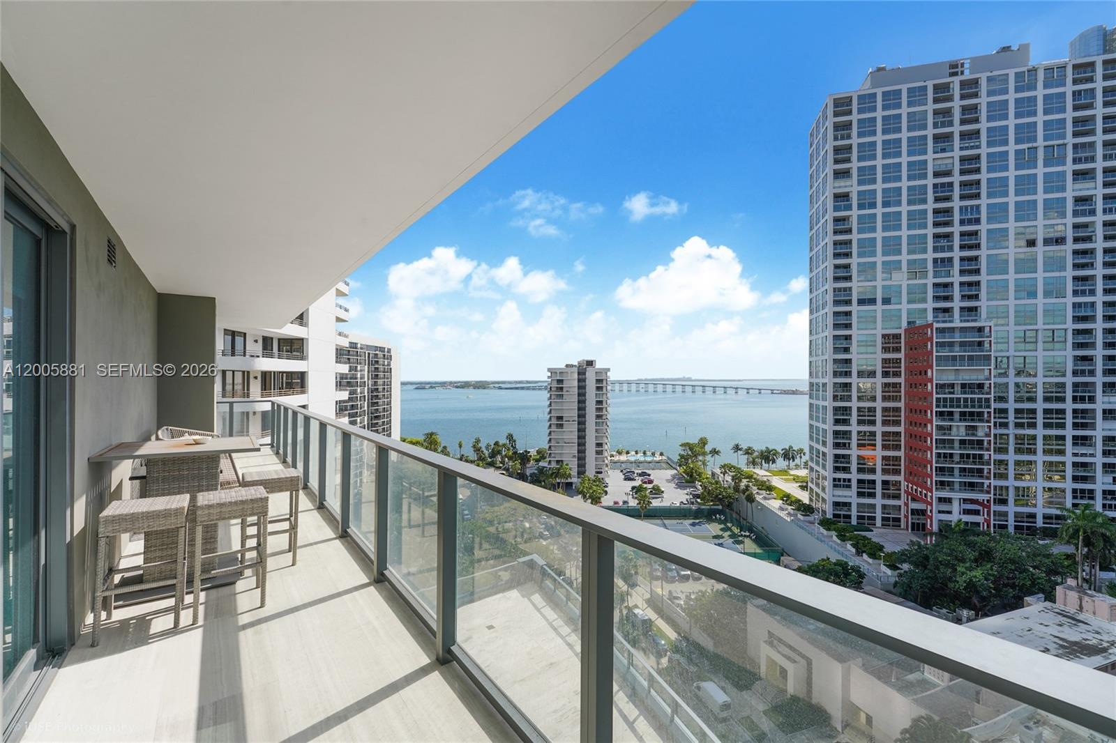 A balcony with outdoor seating overlooks a bay, a bridge, and nearby high-rise buildings — 1451 Brickell Ave # 1203, Miami FL 33131, MIAMI, FL