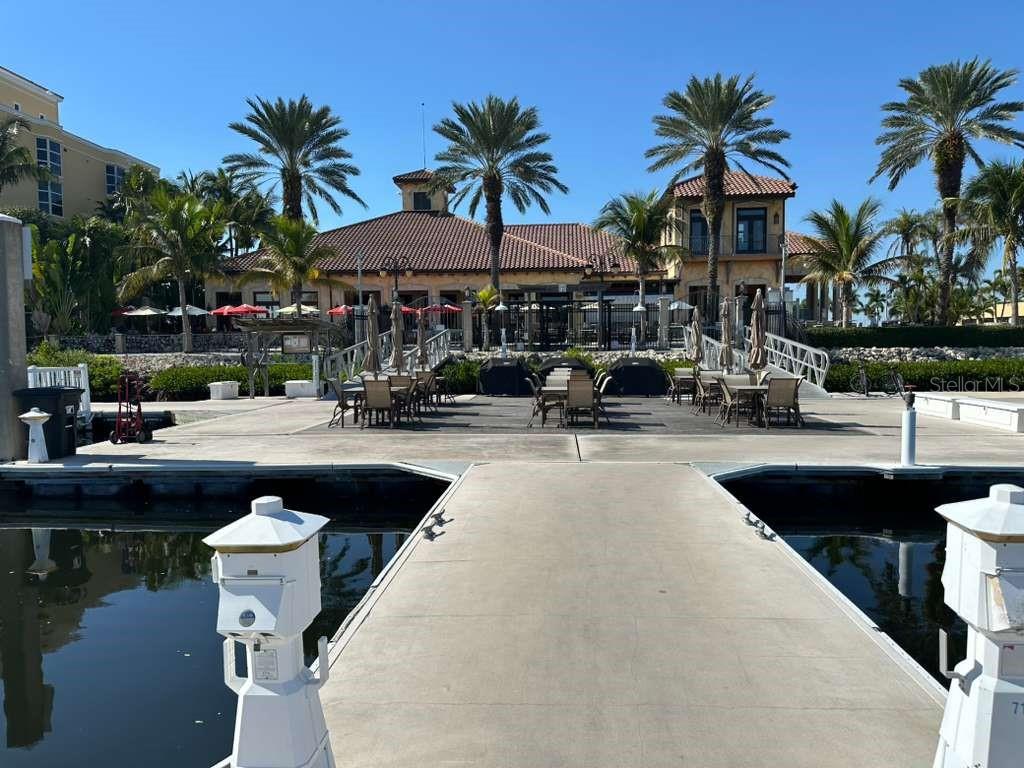 A concrete dock leads toward a waterfront clubhouse with outdoor patio seating and palm trees — 102 RIVIERA DUNES WAY UNIT C29, PALMETTO, FL