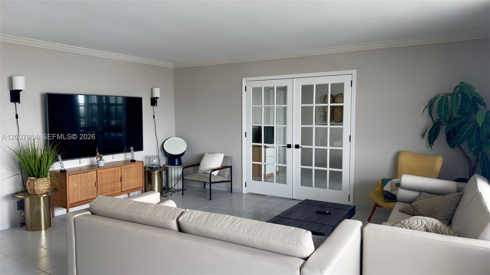 A living room features a large television, white French doors, and light-colored tile flooring — 9225 Collins Ave # 812, Surfside FL 33154, SURFSIDE, FL