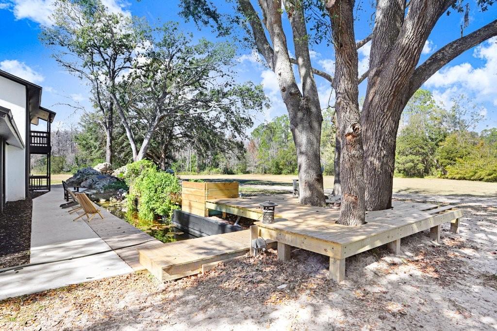 A wooden deck surrounds large trees next to a small pond and a house exterior — 17308 TOBACCO ROAD, LUTZ, FL