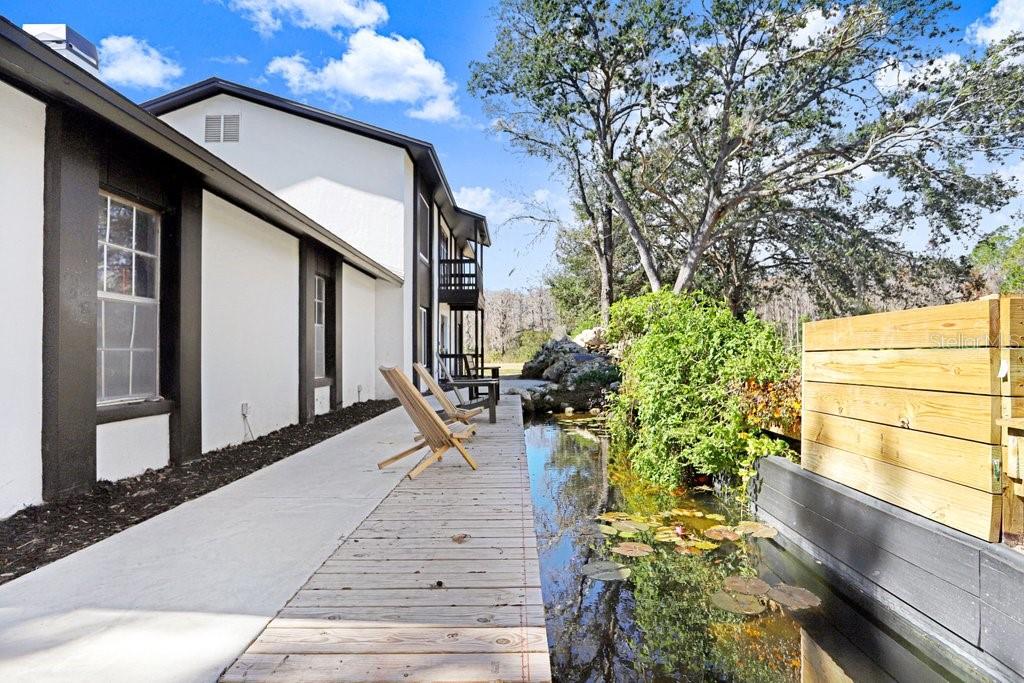 A wooden deck with chairs sits beside a small pond next to a white building — 17308 TOBACCO ROAD, LUTZ, FL