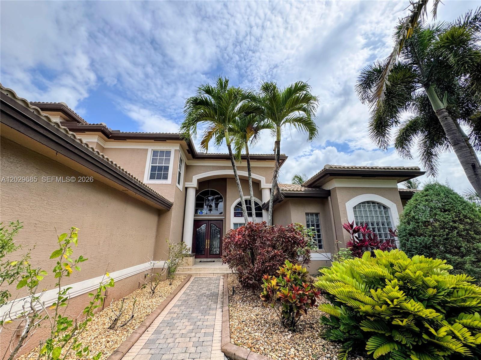 A tan stucco house with a paved walkway, palm trees, and a double front door — 10445 Canterbury Ct, Davie FL 33328, DAVIE, FL