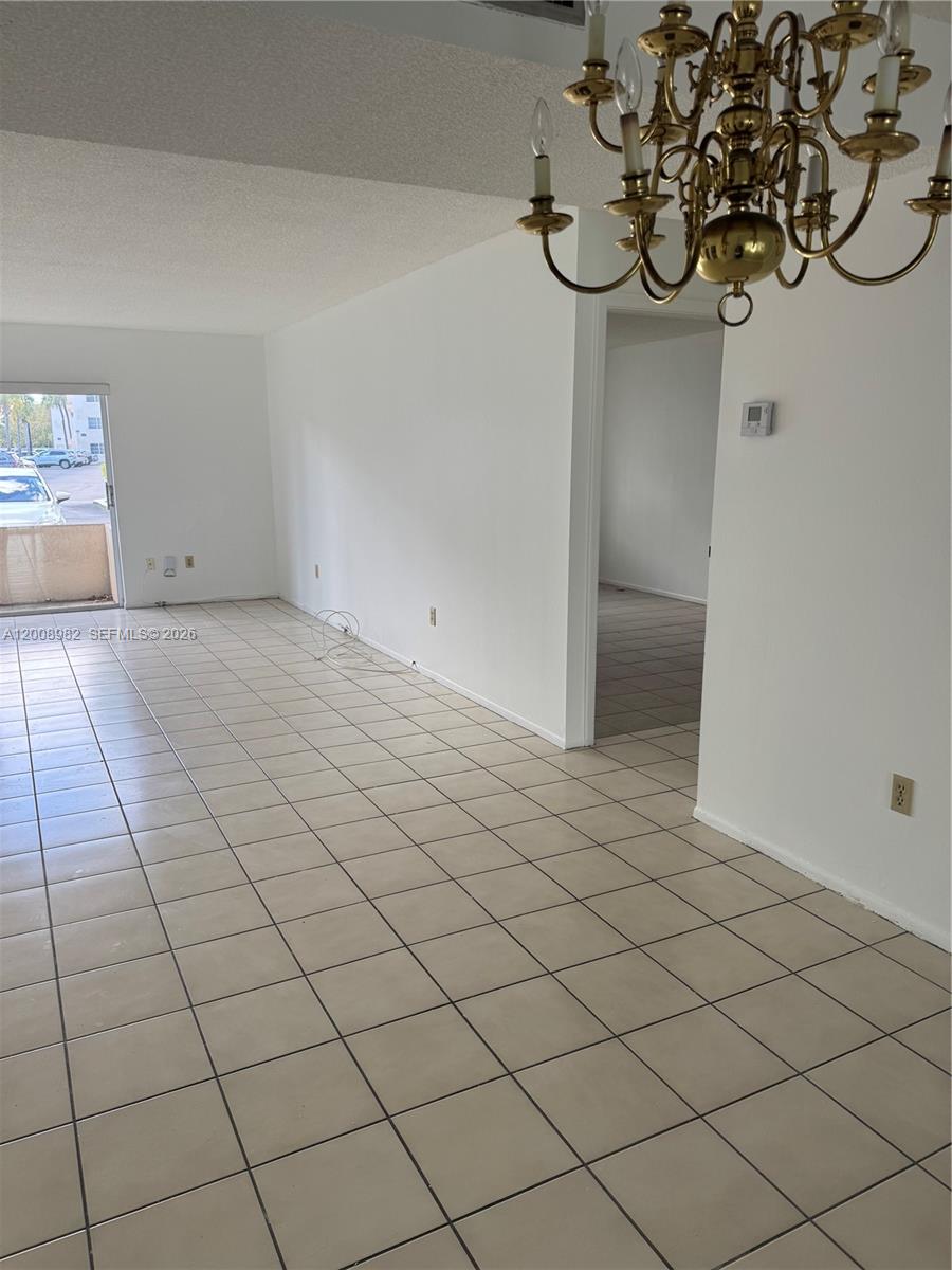 An empty room features white walls, beige tile flooring, and a brass chandelier — 7747 SW 86th St # 105, Miami FL 33143, MIAMI, FL