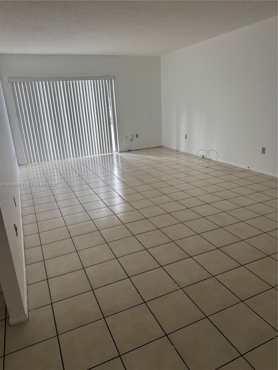 An empty living room features white walls, beige tile flooring, and vertical window blinds — 7747 SW 86th St # 105, Miami FL 33143, MIAMI, FL