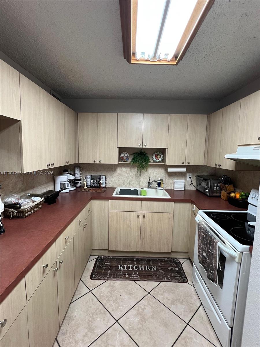 A kitchen features light wood cabinets, red countertops, white appliances, and a tiled floor — 1900 W 68th St # F104, Hialeah FL 33014, HIALEAH, FL