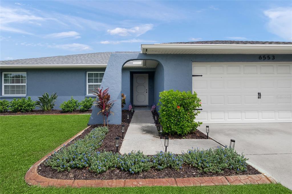 A blue house exterior features a white garage door, a front walkway, and landscaped garden beds — 6553 SAMOA DRIVE, SARASOTA, FL