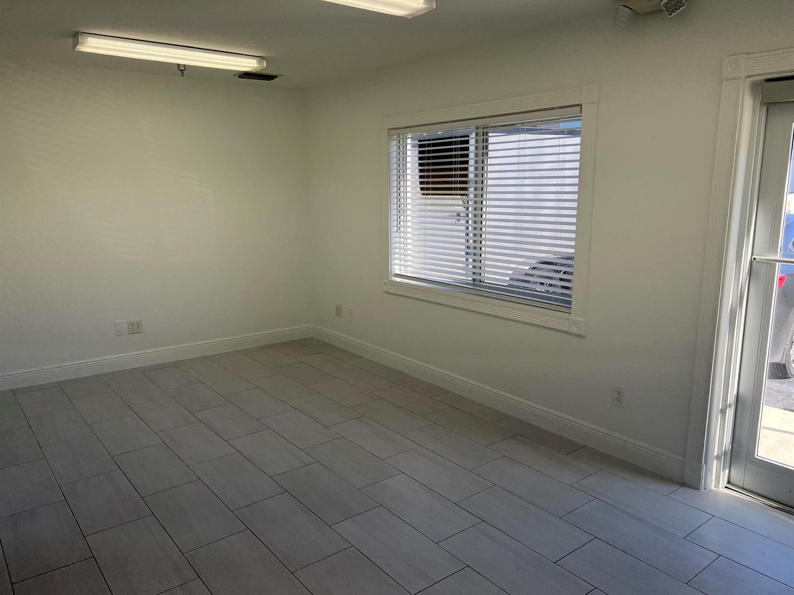 An empty room features white walls, light-colored tile flooring, a window with blinds, and a door — 1312 Commerce Lane # 15b, Jupiter FL 33458, JUPITER, FL