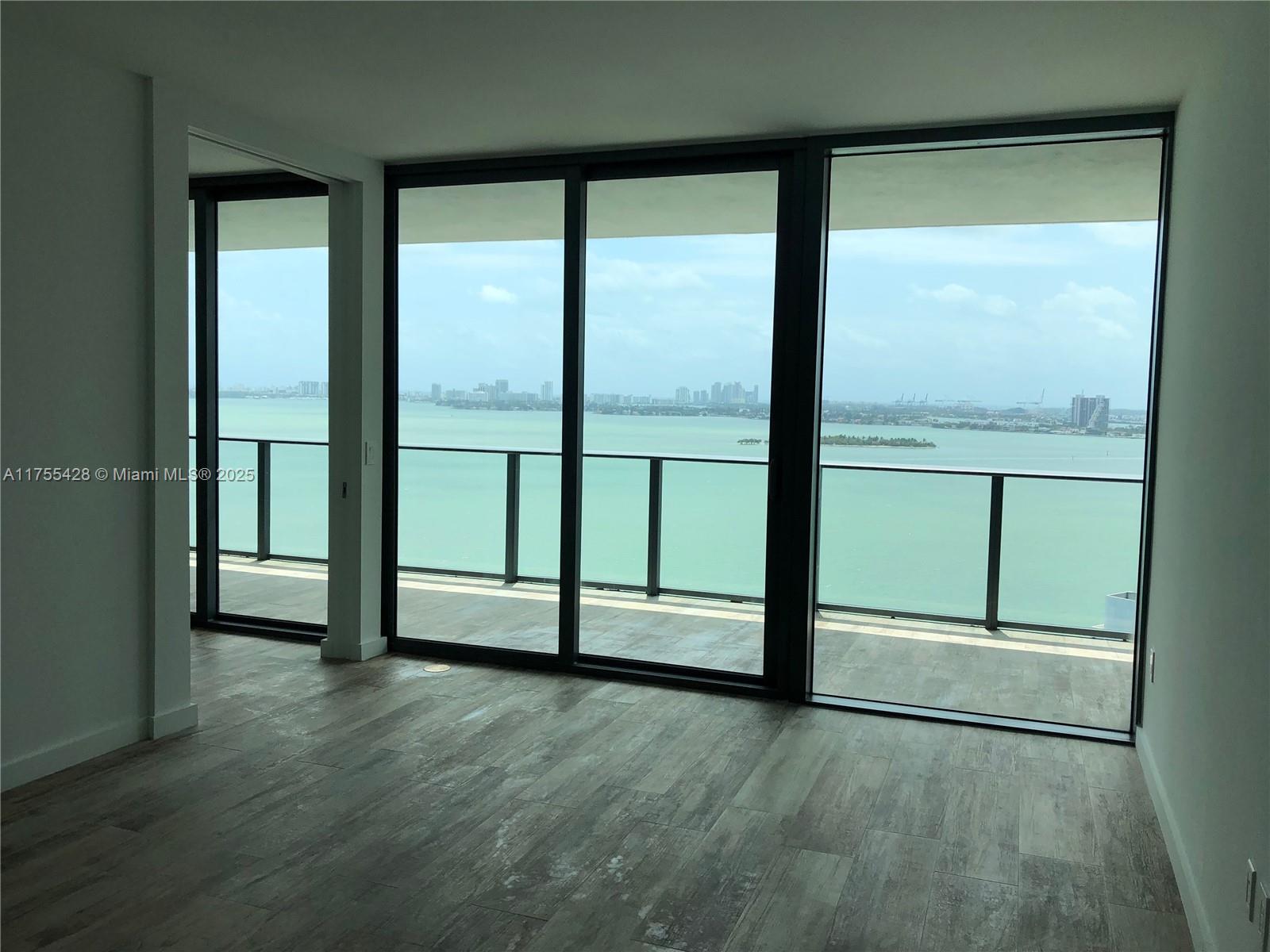 An empty room features wood-look flooring and floor-to-ceiling windows overlooking a bay and city skyline — 650 NE 32nd St # 2006, Miami FL 33137, MIAMI, FL