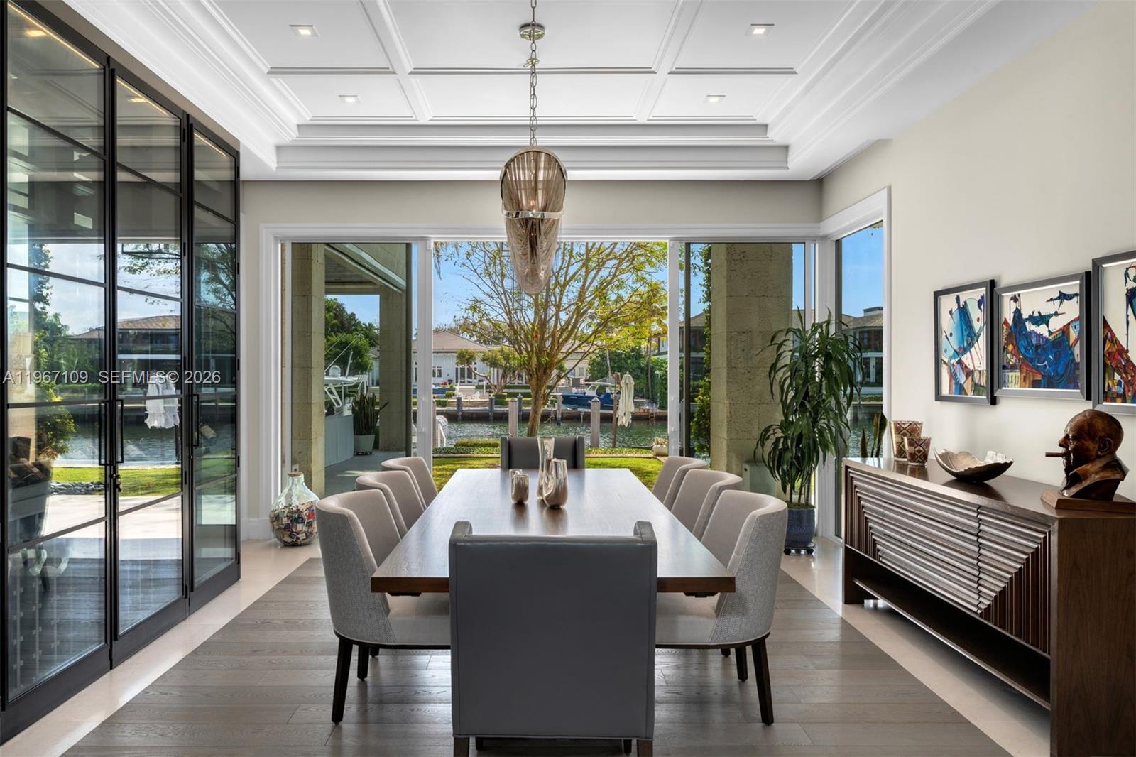 A dining room with a long table, upholstered chairs, and large sliding doors overlooking a canal — 550 Marquesa Dr, Coral Gables FL 33156, CORAL GABLES, FL