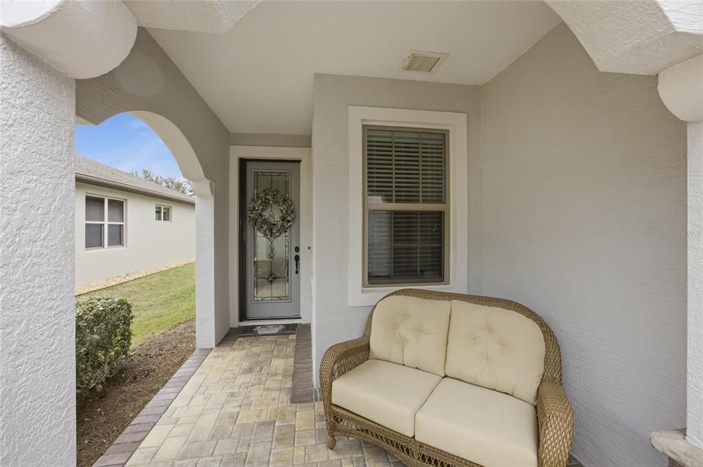 A covered front porch features a wicker loveseat, a glass-paneled front door, and paver flooring — 7639 SW 94TH CIRCLE, OCALA, FL