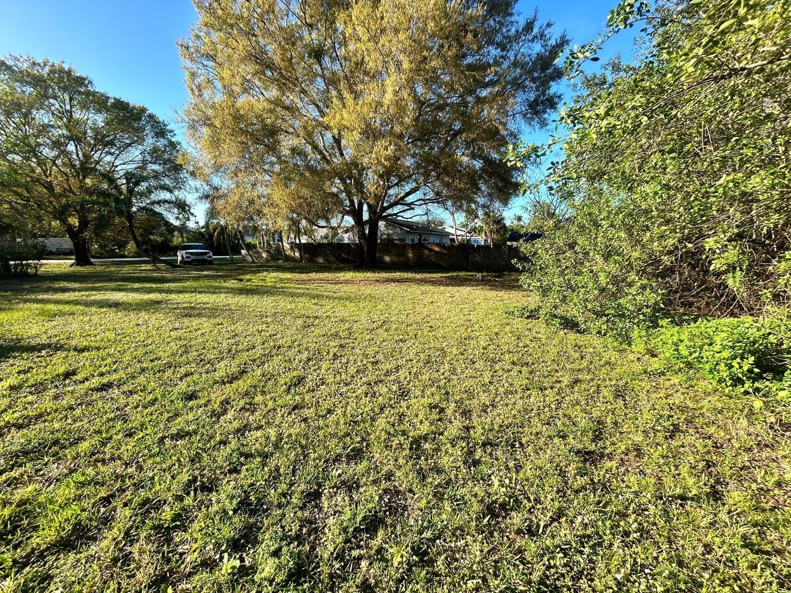 A grassy, open lot features large trees and a wooden fence in the background — 8922 102nd Avenue, Vero Beach FL 32967, VERO BEACH, FL