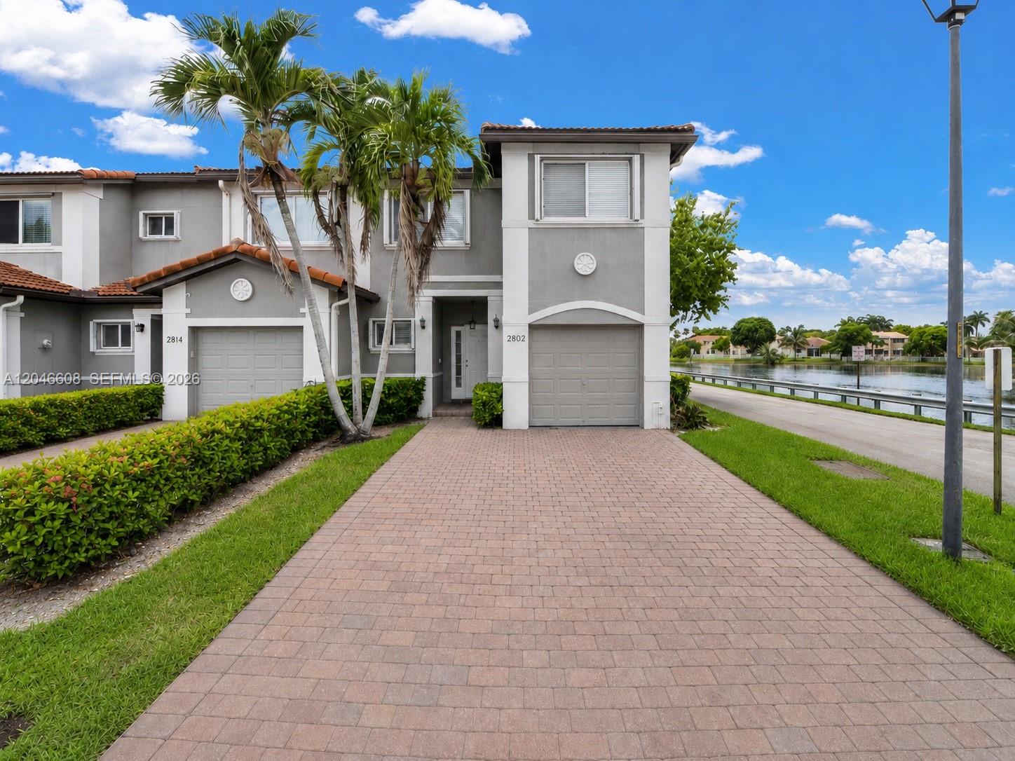 A two-story townhouse with a paved driveway, garage, and palm trees beside a lake — 2802 SW 128th Way # B, Miramar FL 33027, MIRAMAR, FL