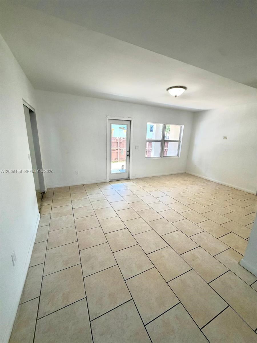 An empty room features white walls, beige tile flooring, a glass door, and a window — MIAMI BEACH, FL