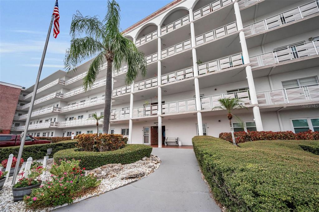 A multi-story apartment building features white balconies, tall support columns, and a landscaped front walkway — 5501 80TH STREET N UNIT 405, ST PETERSBURG, FL