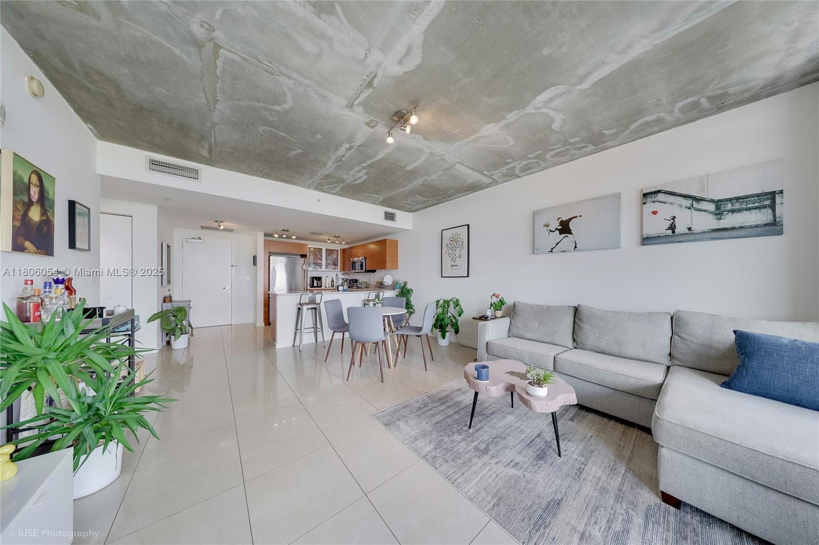 An open-concept living room features a concrete ceiling, tile flooring, and a gray sectional sofa — 3250 NE 1st Ave # 1007, Miami FL 33137, MIAMI, FL