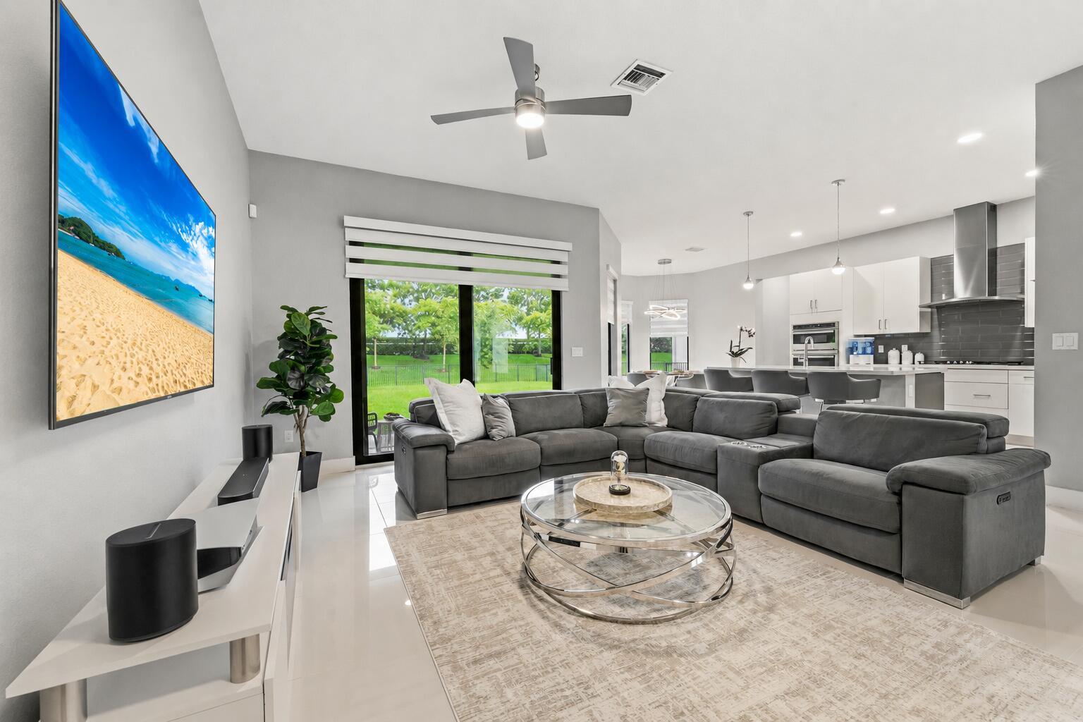 A modern open-concept living room features a gray sectional, glass coffee table, and kitchen area — 10555 Waves Way, Parkland FL 33076, PARKLAND, FL