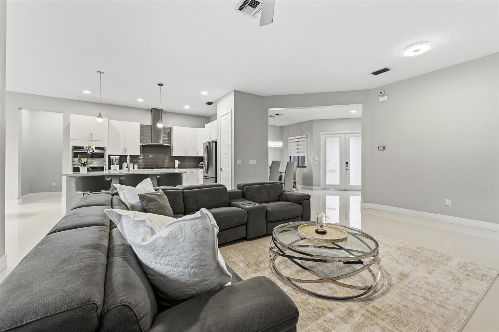 A modern open-concept living room features a large gray sectional sofa and glass coffee table — 10555 Waves Way, Parkland FL 33076, PARKLAND, FL