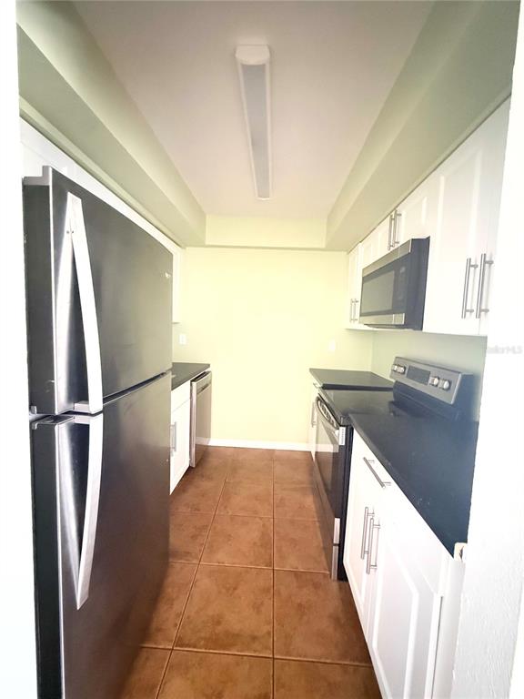 A galley kitchen features white cabinets, black countertops, stainless steel appliances, and brown tile flooring — 1000 LAKE OF THE WOODS BOULEVARD UNIT 101B, CASSELBERRY, FL