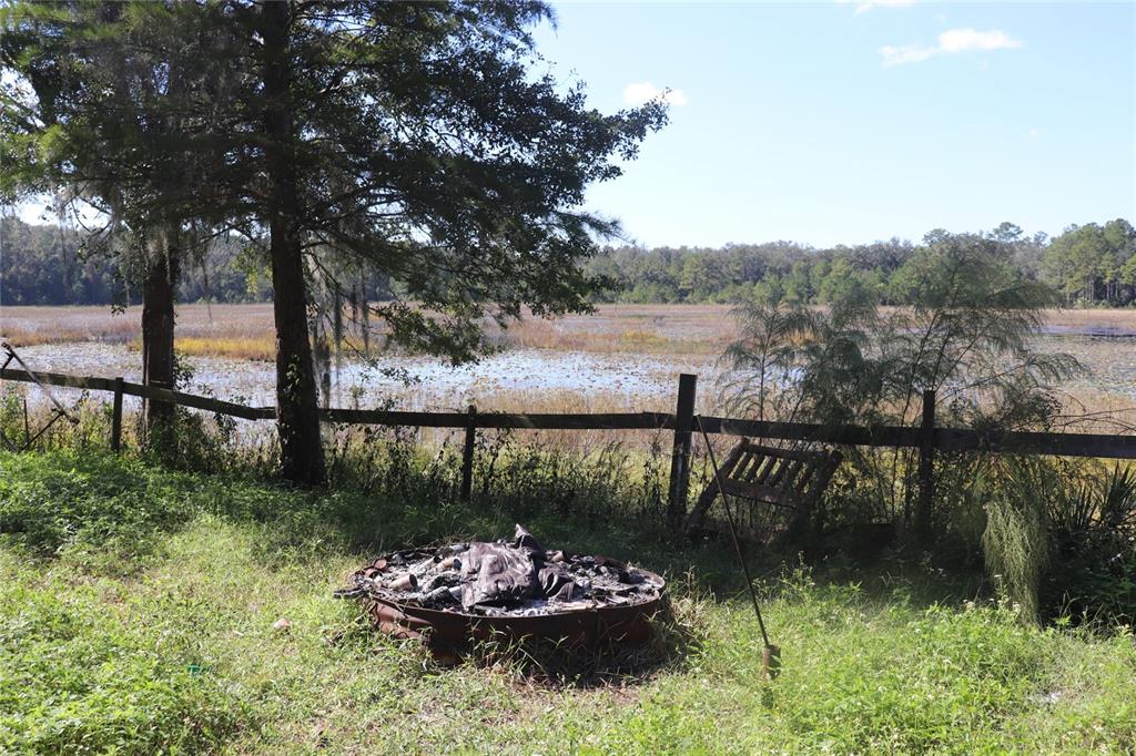 A fire pit sits in a grassy yard before a wooden fence and a marshy lake — 14150 NE 110TH AVENUE ROAD, FORT MC COY, FL