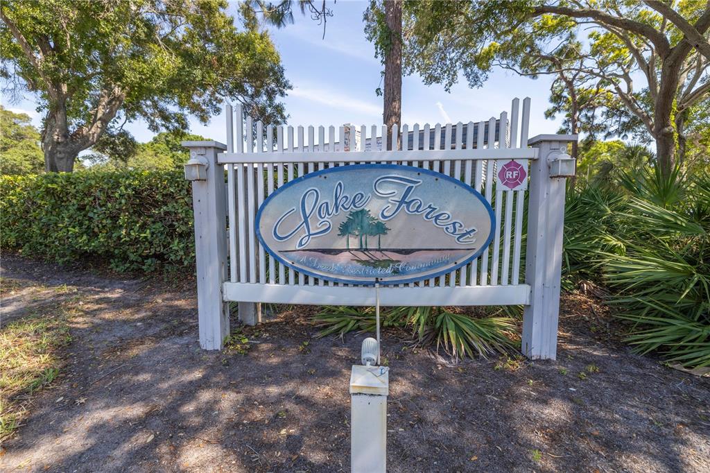 A white picket fence sign for Lake Forest stands outdoors surrounded by trees and bushes — 6462 92ND PLACE N UNIT 803, PINELLAS PARK, FL