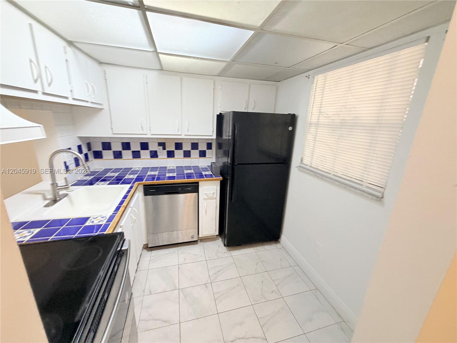 A kitchen features white cabinets, blue-and-white tiled countertops, a black refrigerator, and white flooring — 2800 NW 56th Ave # E201, Lauderhill FL 33313, LAUDERHILL, FL