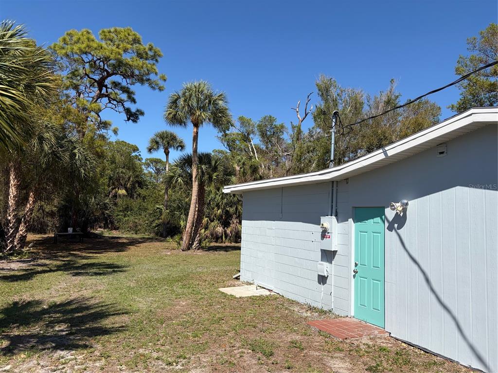 A light blue exterior wall with a teal door stands beside a grassy, wooded yard — 201 PALMETTO ROAD, NOKOMIS, FL
