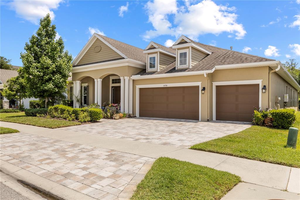 A tan house with a three-car garage and two dormer windows under a blue sky — 16726 COURTYARD LOOP, LAND O LAKES, FL