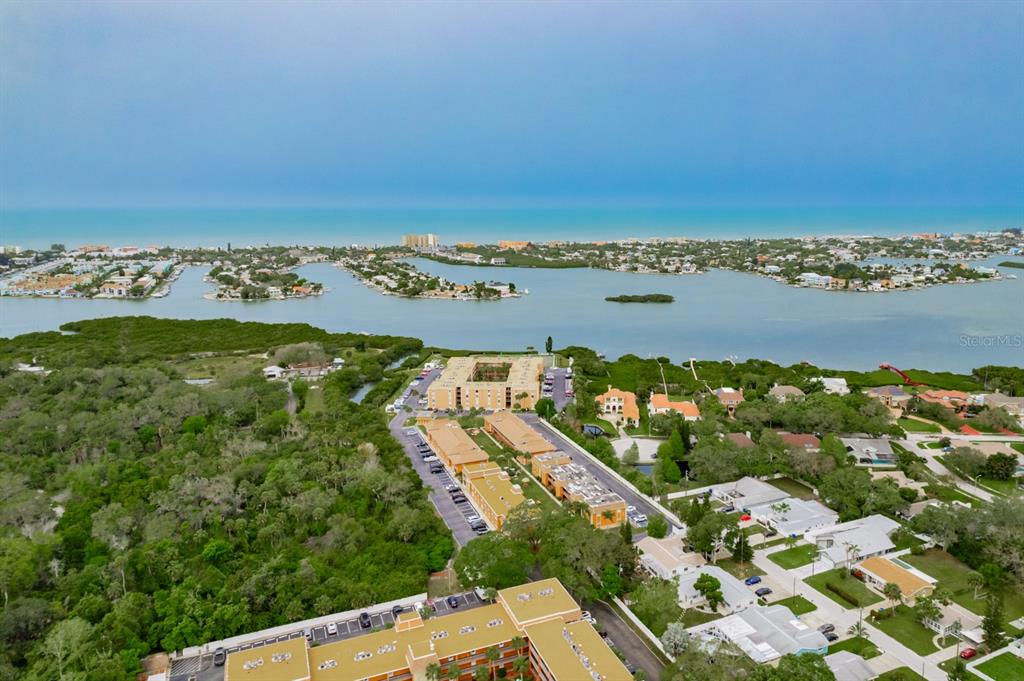 An aerial view shows apartment buildings surrounded by lush greenery, a bay, and the ocean — 12760 INDIAN ROCKS ROAD UNIT 510, LARGO, FL