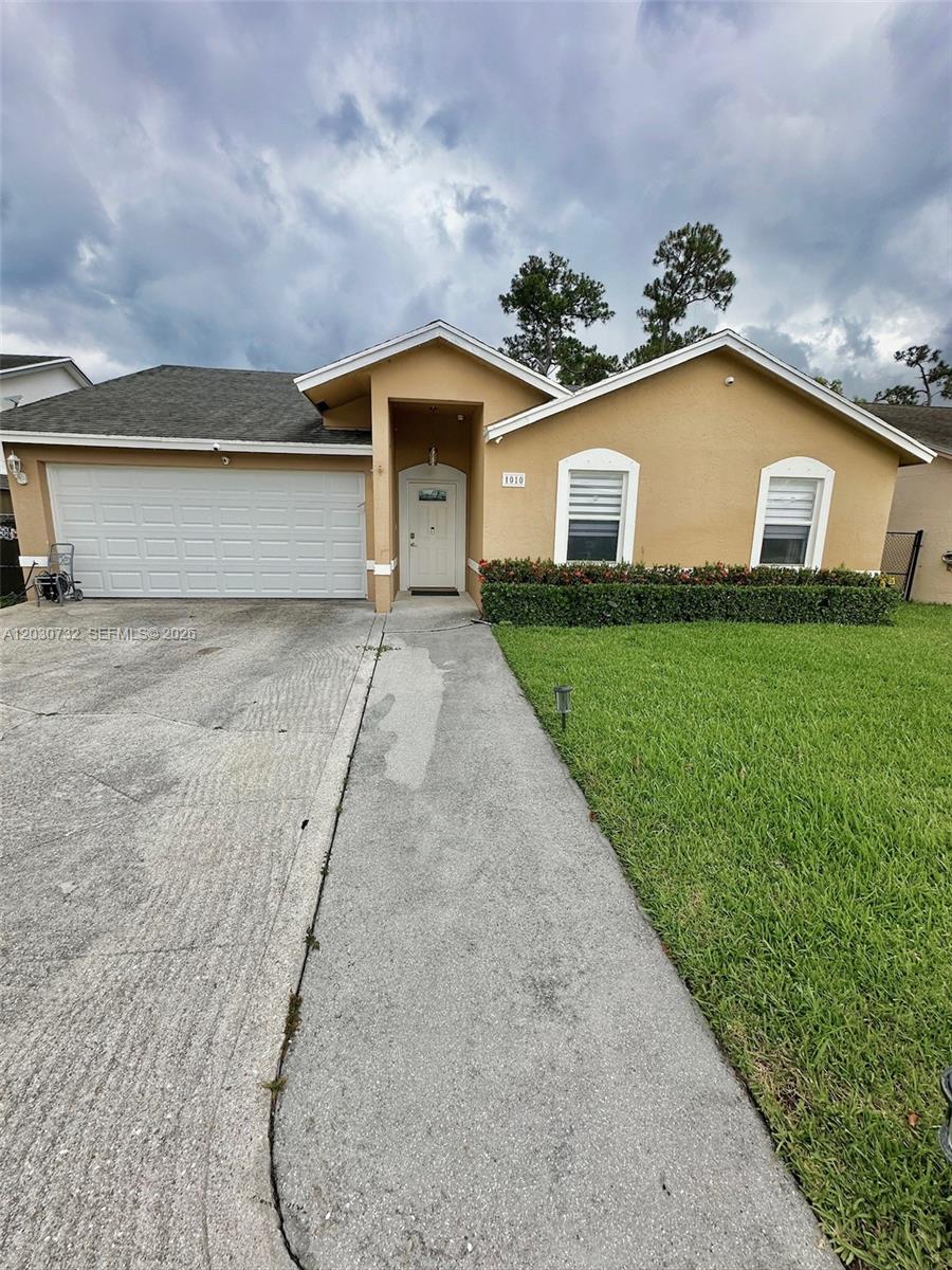 A single-story tan house features a white garage door, a front walkway, and green lawn — 1010 Salmon Isle, Green Acres FL 33413, GREEN ACRES, FL