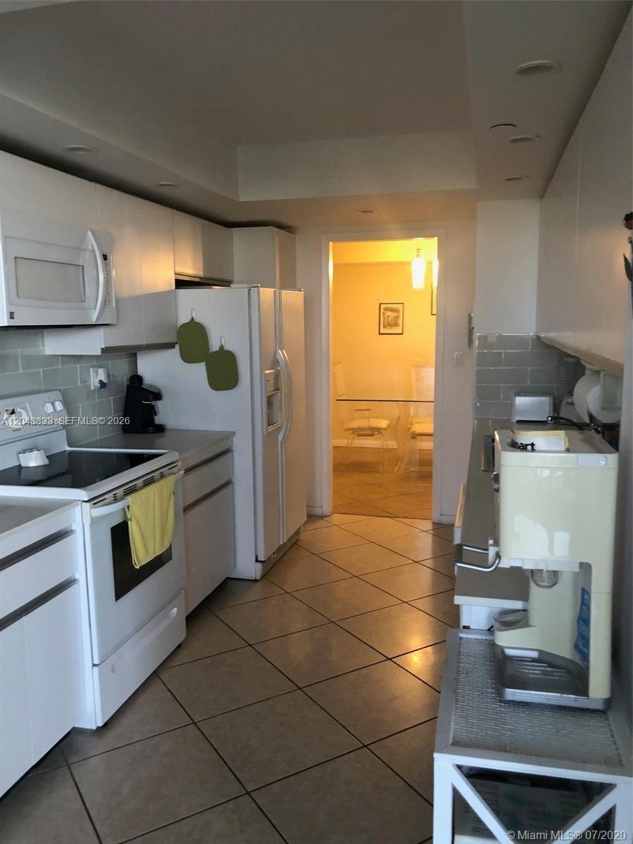 A kitchen with white cabinets, white appliances, and light-colored tile flooring leads to a dining area — 2000 Towerside Ter # 905, Miami FL 33138, MIAMI, FL
