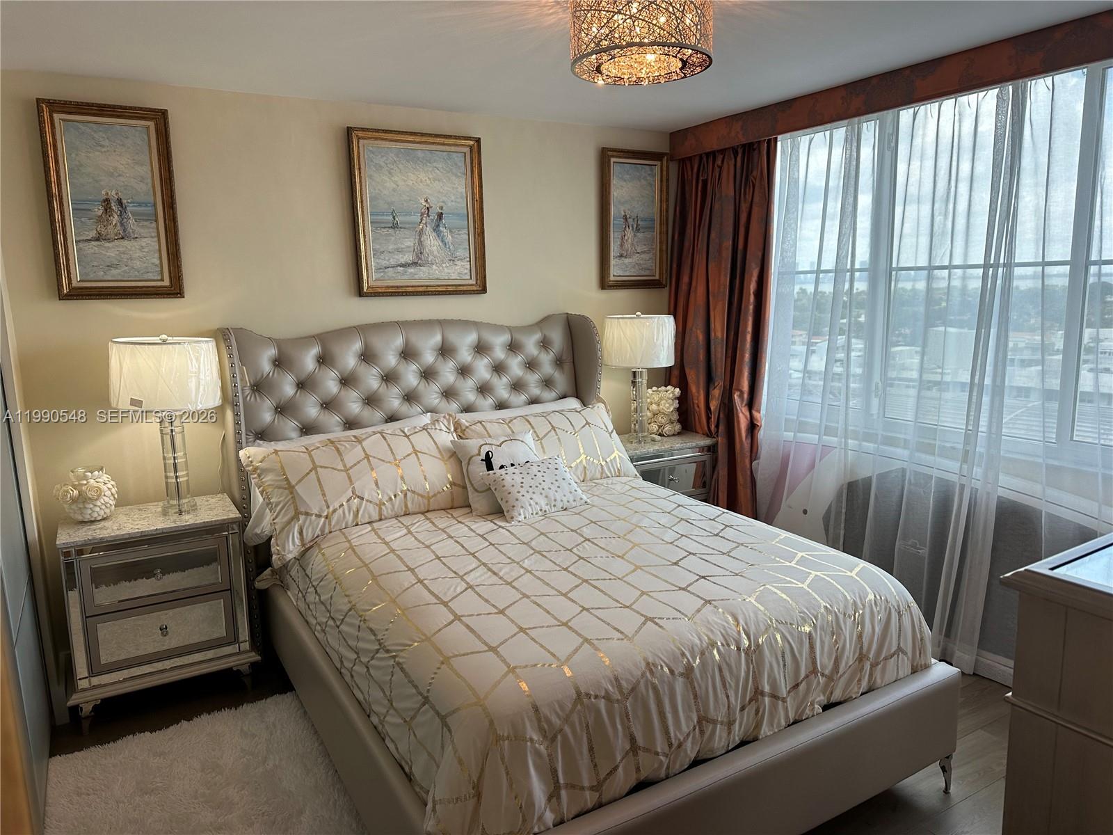A bedroom features a tufted headboard, mirrored nightstands, gold-patterned bedding, and a large window — 6039 Collins Ave # 1431, Miami Beach FL 33140, MIAMI BEACH, FL