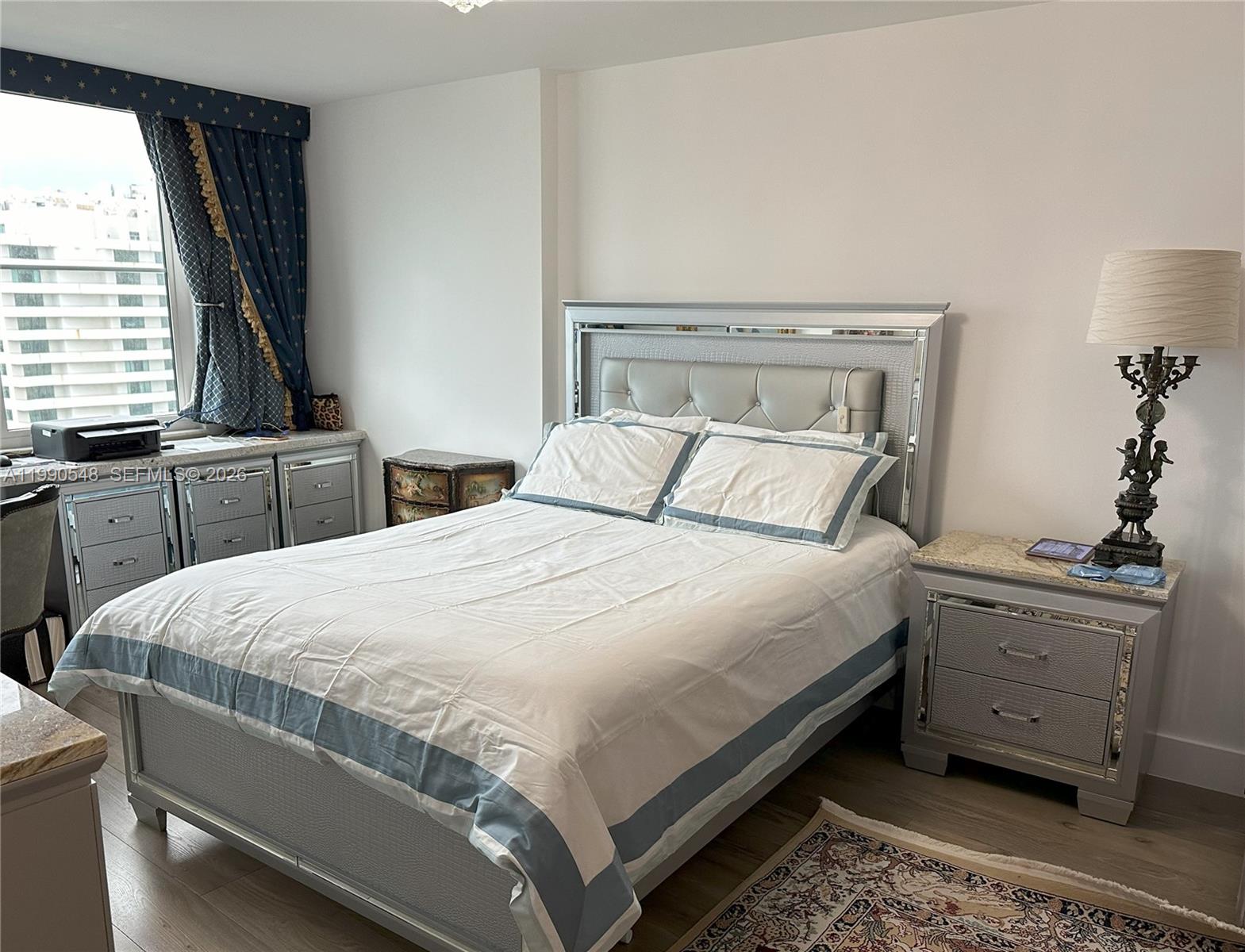 A bedroom features a mirrored bed frame, matching nightstand, and blue patterned curtains by a window — 6039 Collins Ave # 1431, Miami Beach FL 33140, MIAMI BEACH, FL